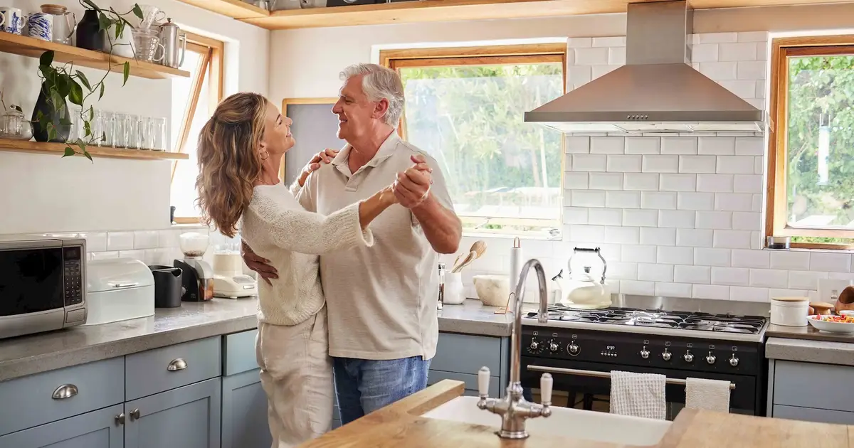 Older couple dancing in sunlit kitchen.