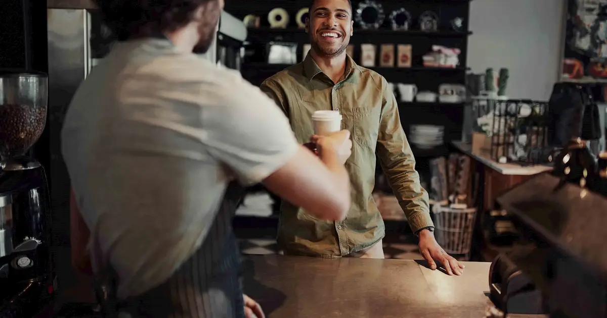 A barista hands a cup of coffee to a smiling customer across the counter in a cozy, dimly lit café. The customer is wearing a green button-up shirt, and the background features coffee beans, cups, and café decor.