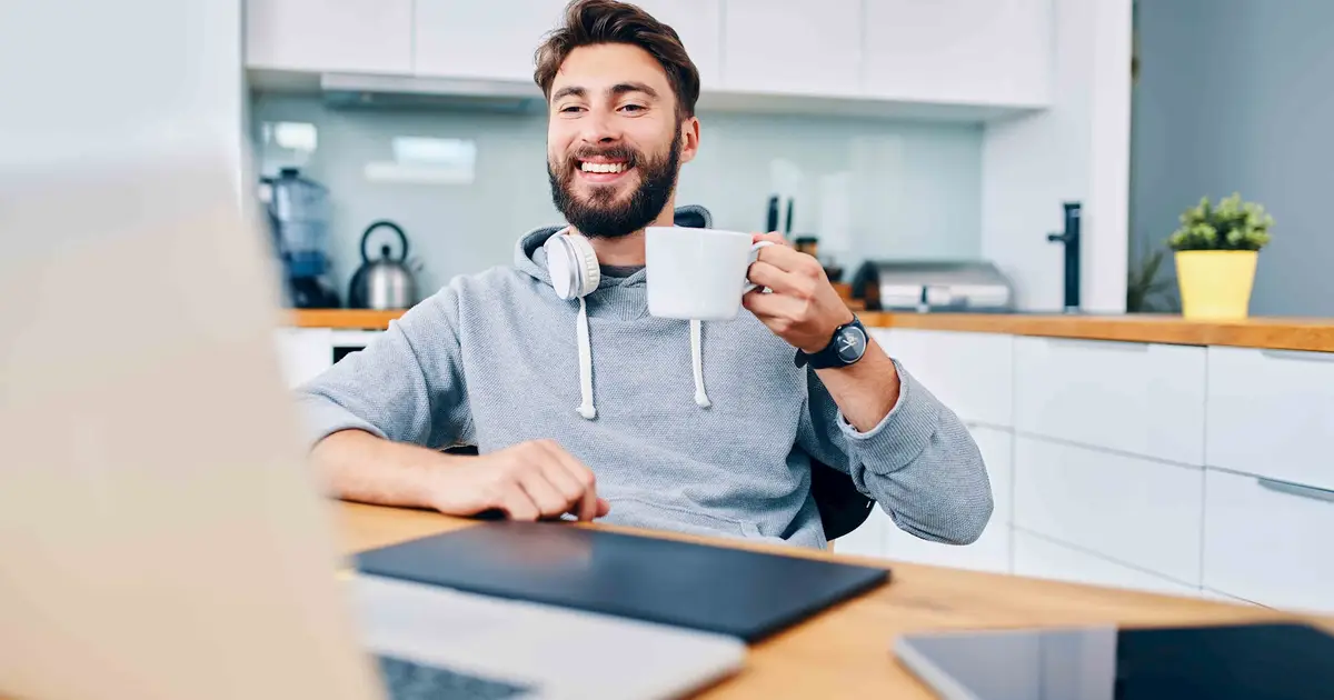 Smiling man with coffee mug works on his laptop in a kitchen.