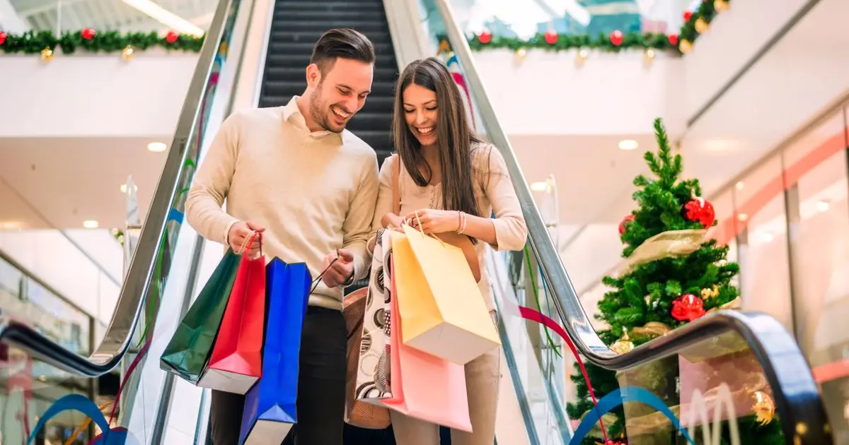 Couple smiling, carrying shopping bags, with holiday decorations and tree.