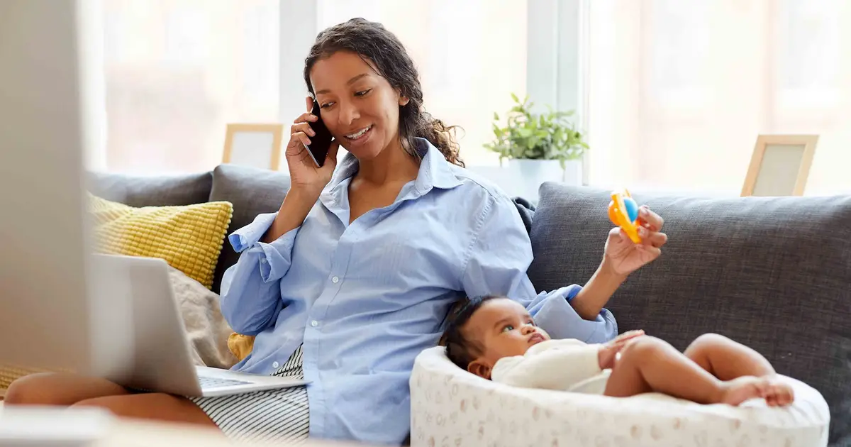 Woman multitasking with baby, phone, and laptop.