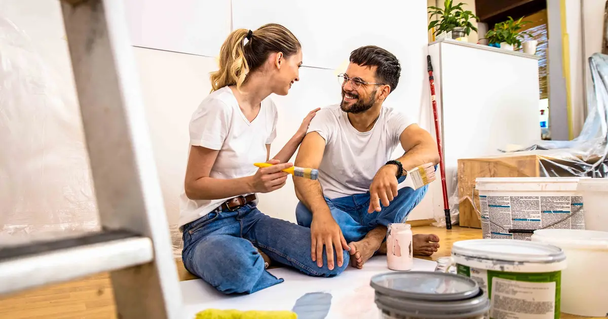 A smiling couple sits on the floor of their home, dressed casually in jeans and white shirts, surrounded by paint supplies. They are engaged in a light-hearted moment while taking a break from painting, with paint cans, brushes, and rollers nearby. The woman holds a paintbrush, gently touching the man's arm, and they share a warm, playful look. The room is in the process of being painted, with protective plastic covering some furniture in the background.