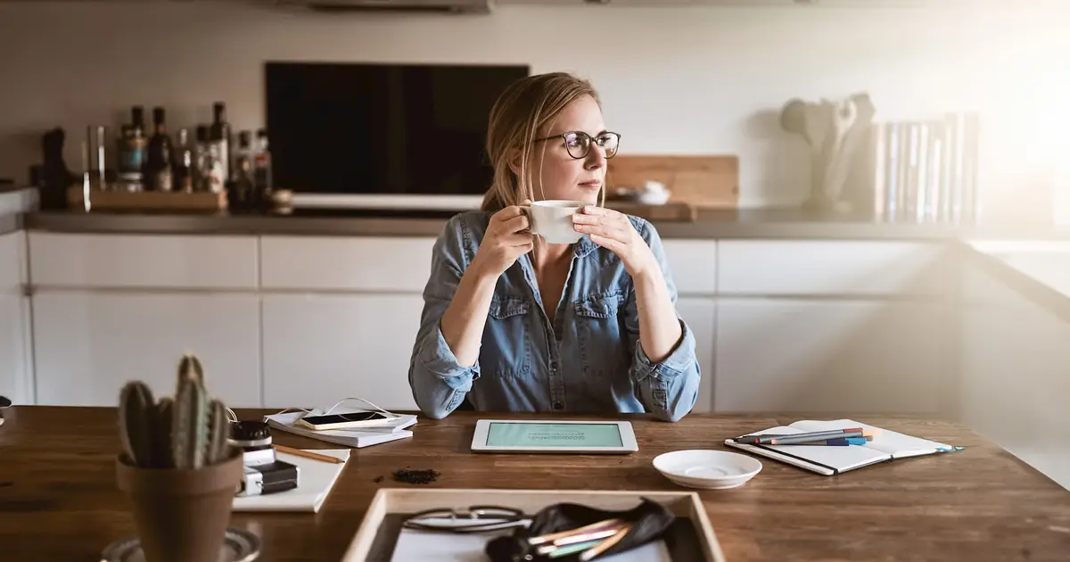 A woman in a casual denim shirt sits at a wooden table, sipping coffee while gazing thoughtfully into the distance. Her desk is cluttered with a tablet, notebooks, and pens.