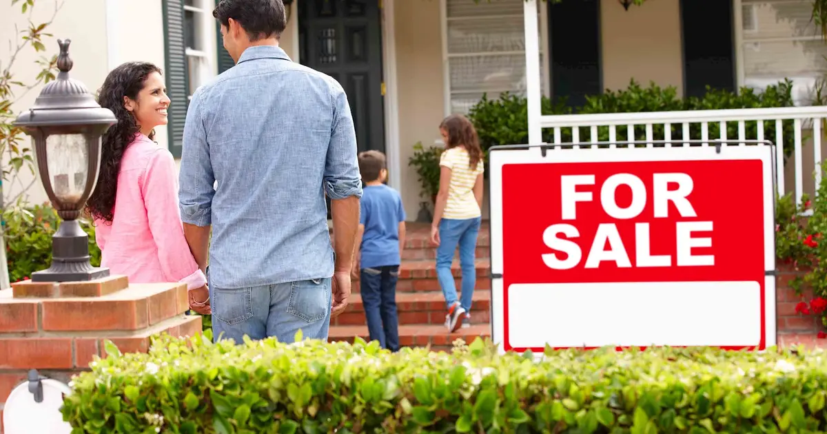 A family of four stands in front of a house with a large 'For Sale' sign, looking excited as they walk towards the entrance. The parents, holding hands, smile at each other while their two children, a boy and a girl, approach the front steps. The house has greenery around the porch, giving it a welcoming and homey feel.