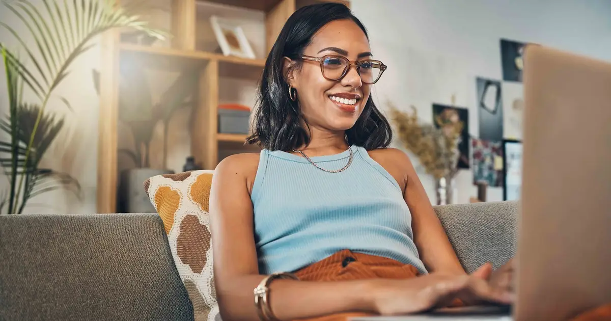 A woman sits on a couch in a cozy living room, smiling while working on a laptop. She is wearing glasses, a light blue sleeveless top, and a necklace. The background features wooden shelves, decorative plants, and a soft pillow with geometric patterns.
