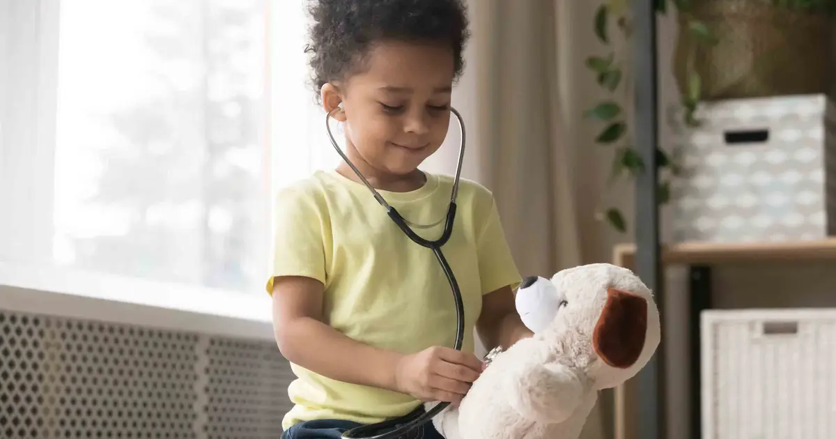 Child in yellow shirt plays doctor with stuffed animal, smiling.