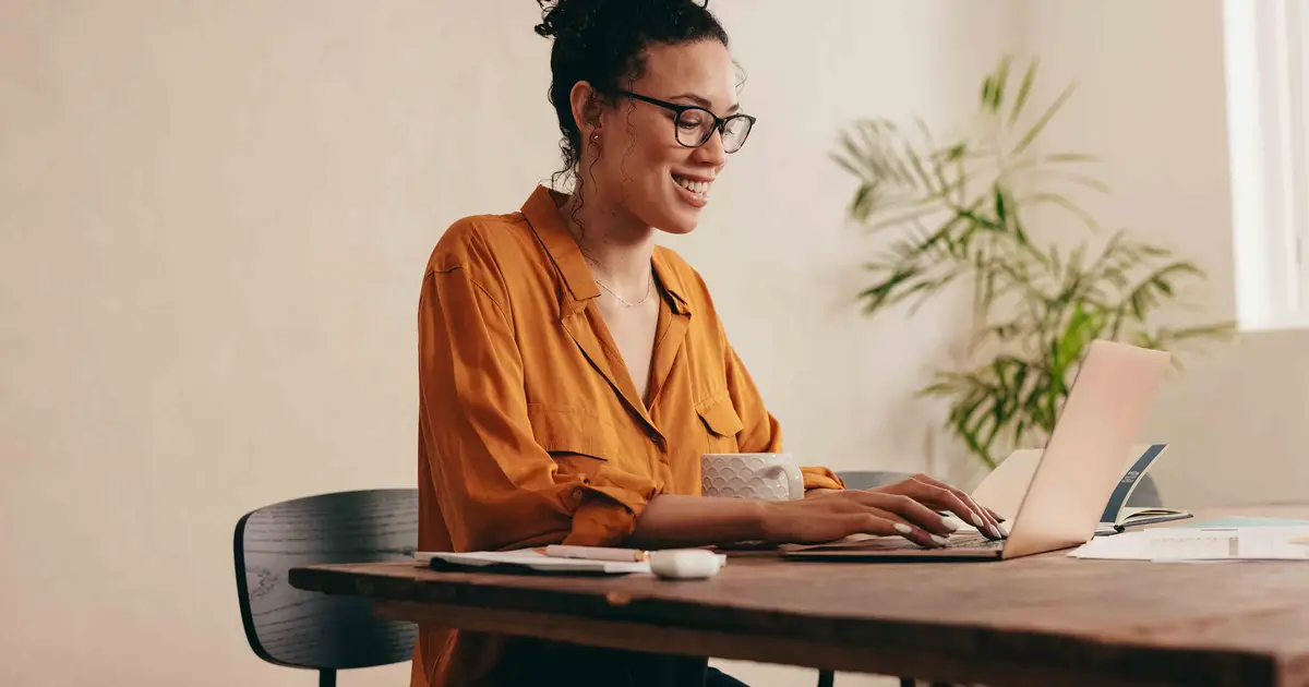 Woman in orange shirt smiles while working on laptop at desk.