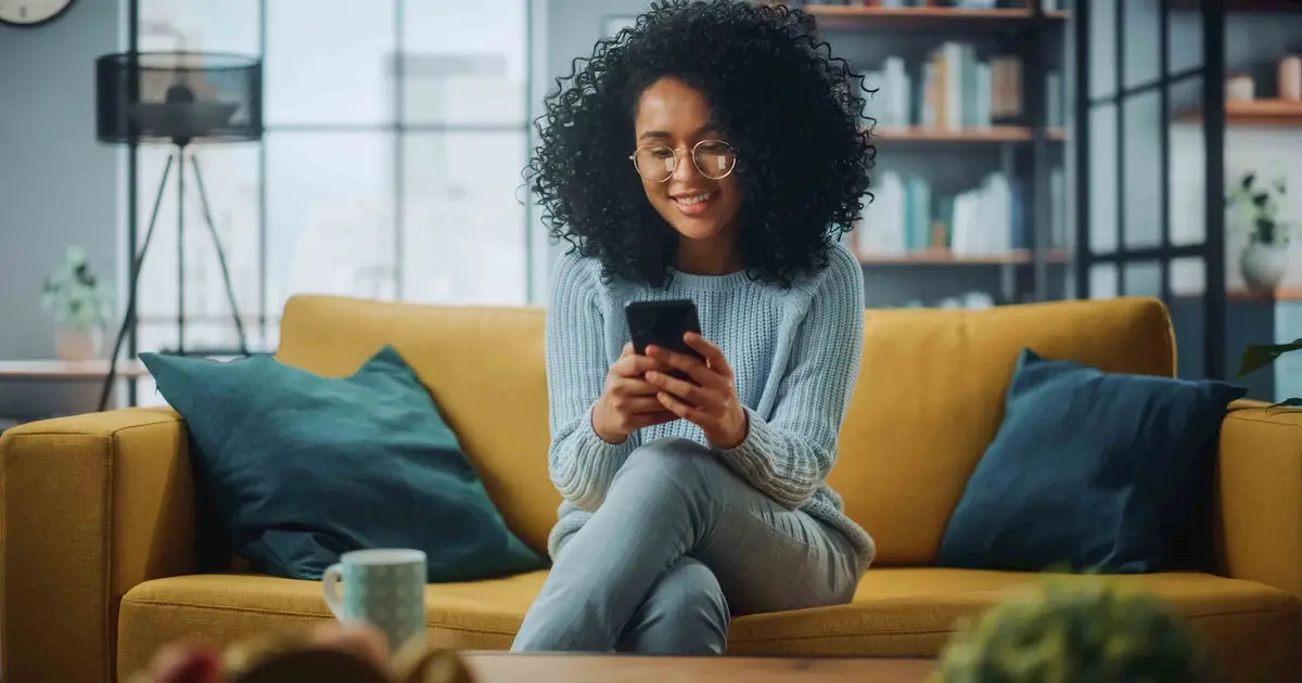 Smiling woman sits on yellow couch, looking at smartphone indoors.