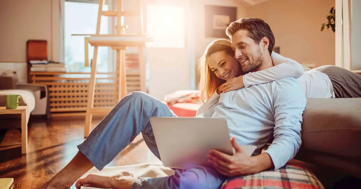 Smiling couple sits together on floor, hugging and viewing laptop screen.