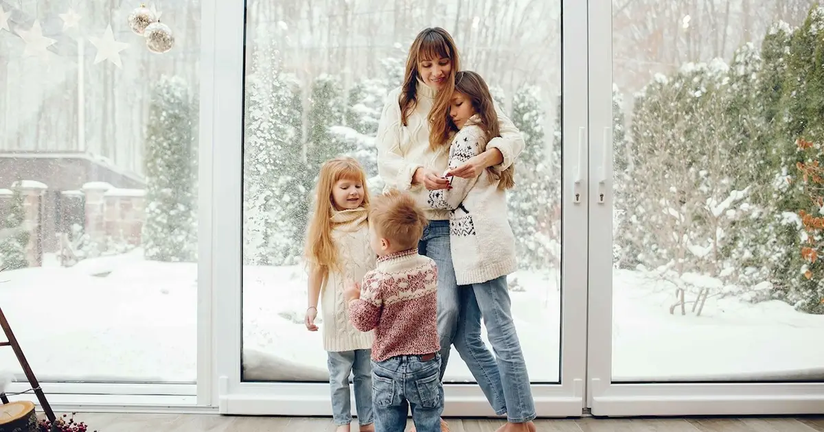 Mother and three kids in sweaters admire snowy view by window.