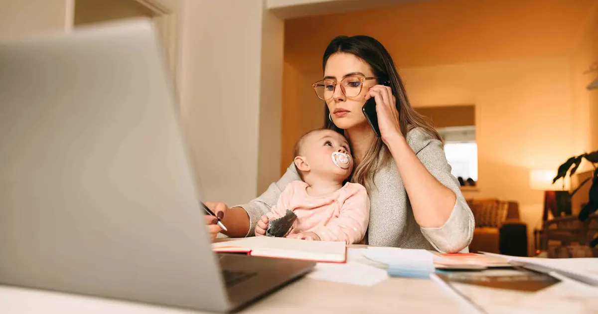 Woman multitasking with baby, phone, and laptop in home office.