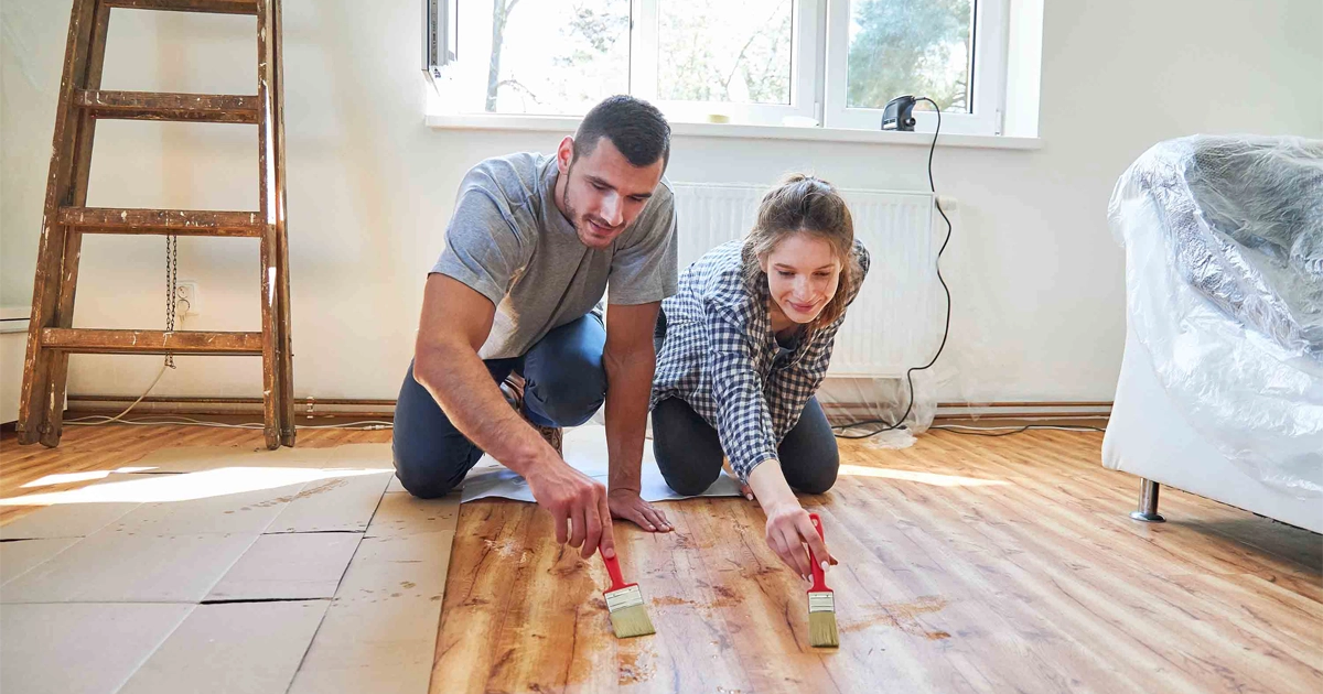 Couple staining hardwood floors during home renovation with ladder and furniture nearby.
