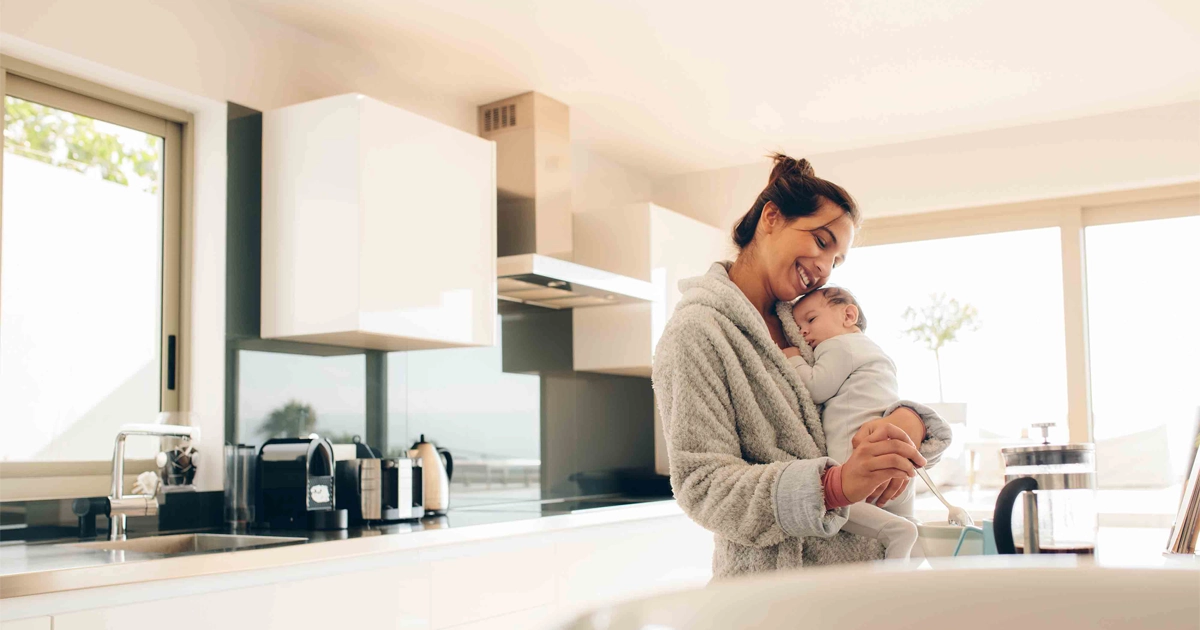 Mother in robe holds sleeping baby while preparing breakfast in sunny kitchen.
