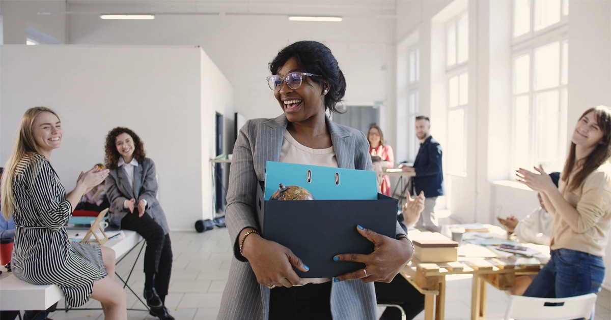 Smiling woman carries box as colleagues applaud.
