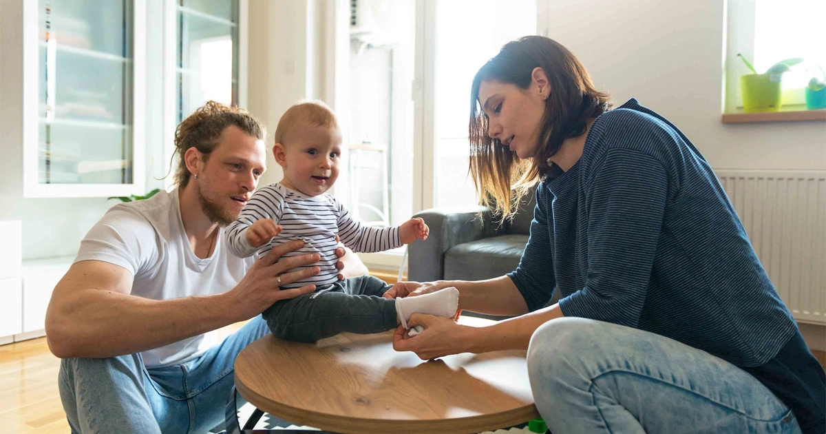 Father holds baby on table while mother puts on baby's socks.