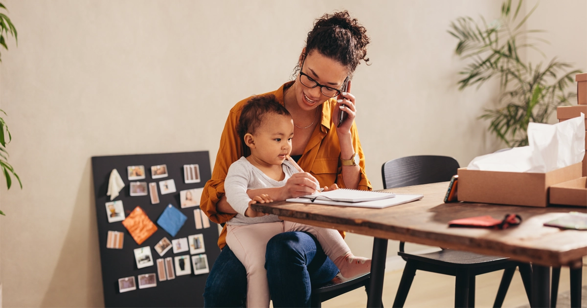Mother works from home with baby on lap holding pen.