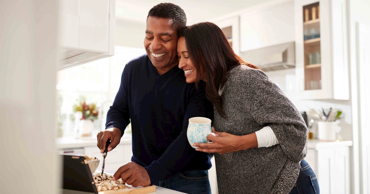 Smiling couple cooking in bright kitchen; one chops mushrooms, one holds mug.