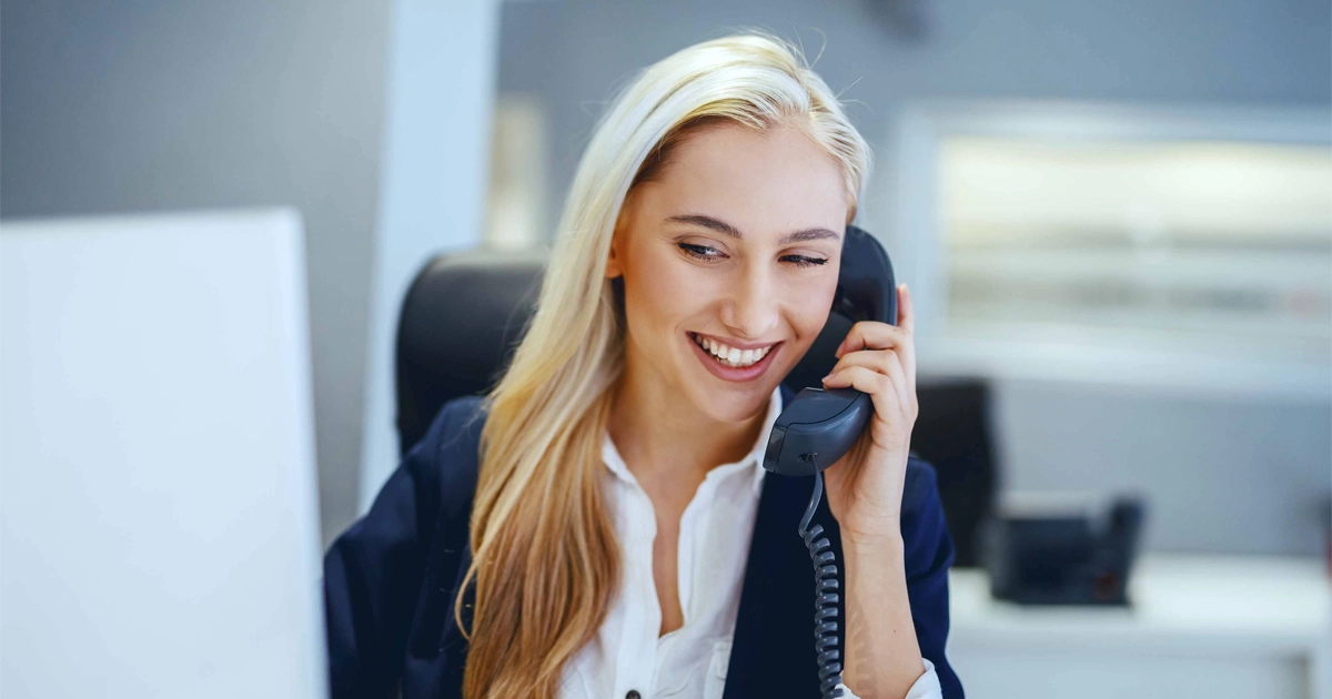 Smiling professional woman in blazer talks on phone at office desk.