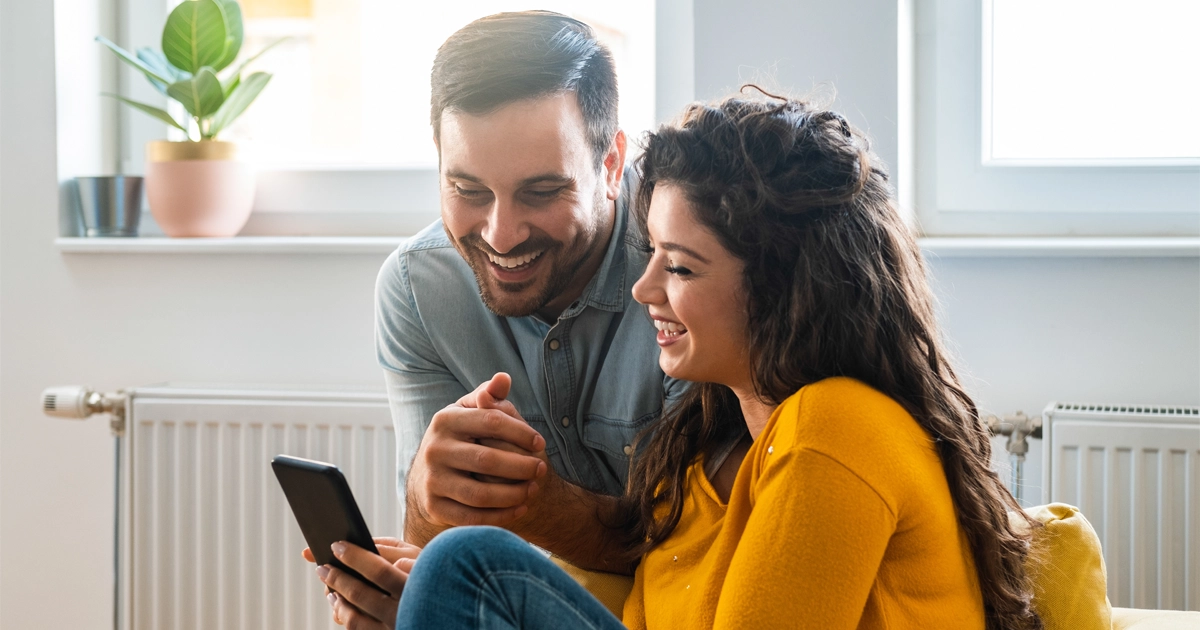 Smiling couple on couch looking at smartphone in cozy, sunlit living room.